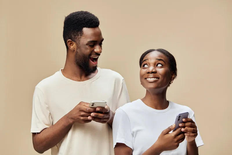 Happy young African couple enjoying time together while using smartphones against a soft beige background, showcasing modern casual clothing and playful emotions