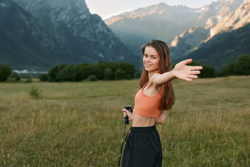 A cheerful woman stands in a meadow with distant mountains, extending her arm toward the camera, smiling warmly to create a bright, energetic outdoor mood.