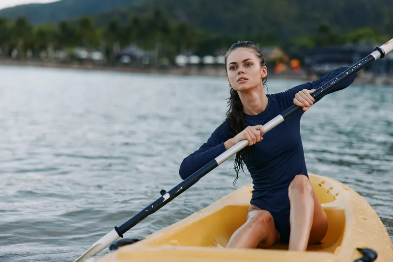 A focused female rower paddles a yellow kayak across calm water, gripping the paddle with determination as a scenic shoreline and hills blur in the background.