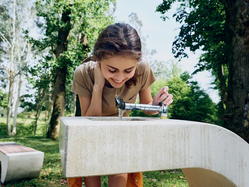 Girl drinking water from a concrete fountain in a sunny park, child leaning over the tap for refreshment, casual summer portrait showing hydration, smile and playful outdoor moment.