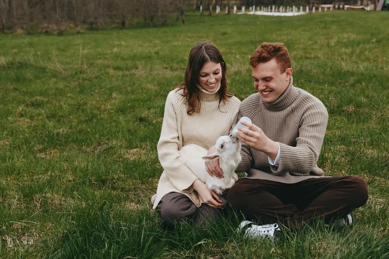 Happy couple outdoors with a fluffy goat kid, sharing a gentle moment on a green field, cozy sweaters, warm smiles, natural light, candid lifestyle scene.
