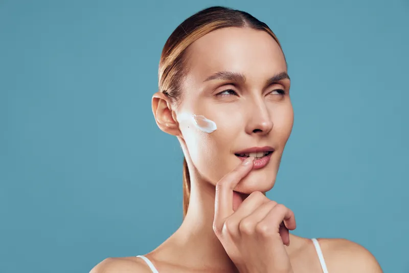 Skincare woman young face with cream on cheek, moisturize beauty spa portrait, closeup on calm expression, soft blue background, gentle finger near lips, care today
