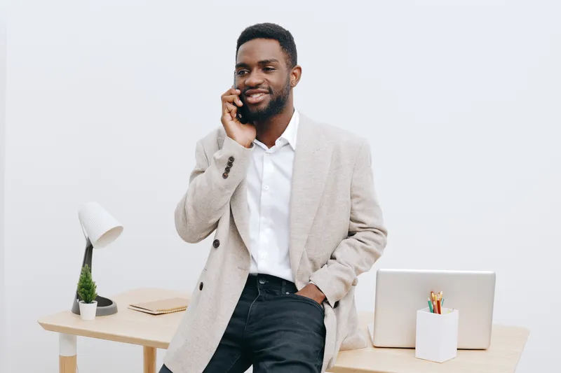 Professional young African-American man in smart casual attire smiling while talking on mobile phone in modern office environment. Business communication, success, confident behavior, coworking space,