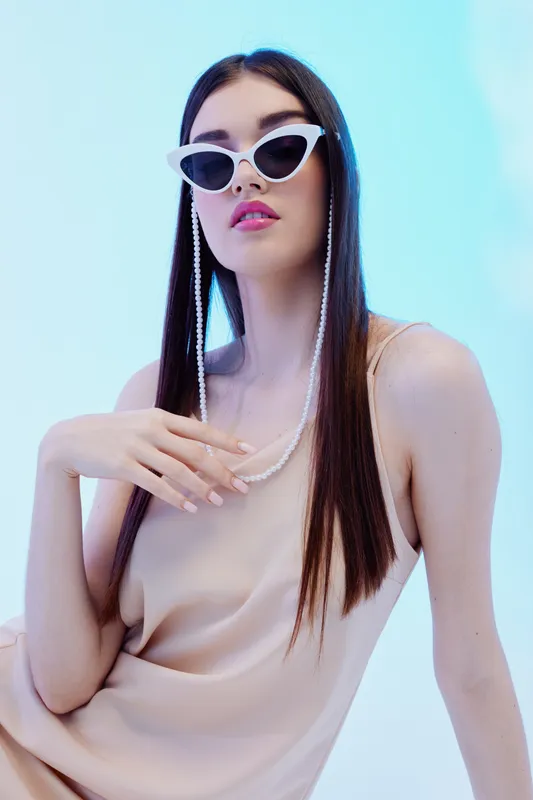 Fashionable woman with long straight hair wearing white cat-eye sunglasses and a beige sleeveless top poses against a soft blue background. Concept of beauty, style, and elegance.