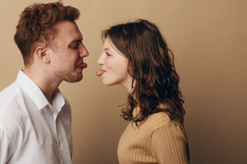 Young man and woman face each other playfully sticking out tongues in casual clothes on beige background. Concept of fun, humor, relationship, and lighthearted interaction between couple.