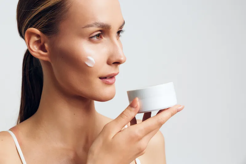 Closeup of a woman applying moisturizer from a jar, soft light highlights flawless skin, calm expression, and a skincare routine moment, natural colors, clean background, gentle touch.