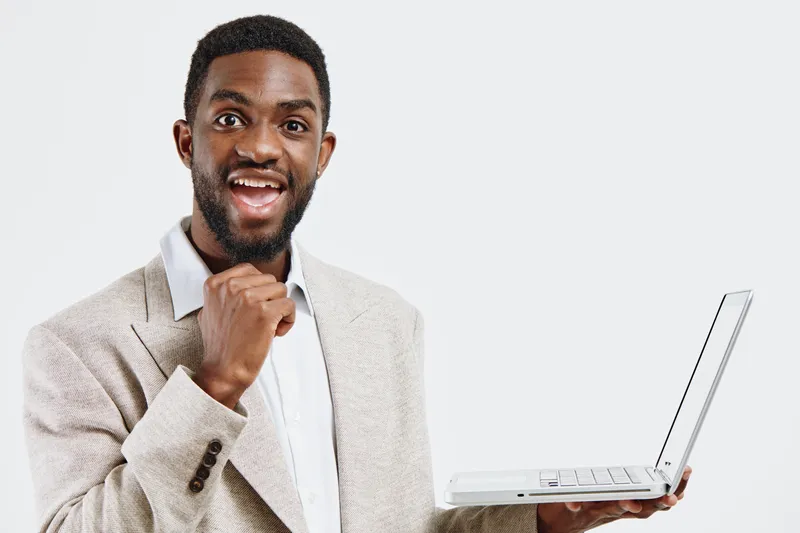 Smiling man holding laptop in hand, wearing formal beige blazer and white shirt, looking excited and confident, studio portrait on plain white background. Business, technology, success, professional, 