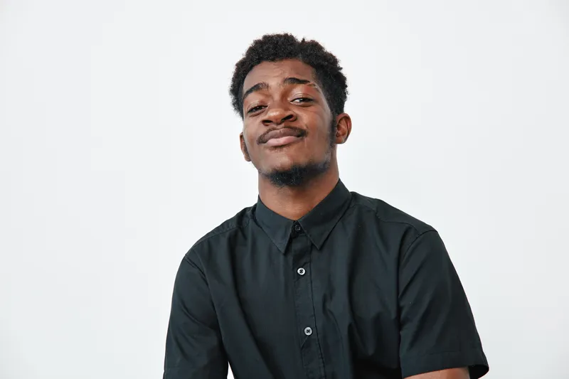 Young African American man with dark skin wearing black shirt posing confidently against white background. Portrait of confident male with short curly hair and casual style.