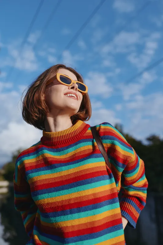 Woman in rainbow sweater and yellow sunglasses smiling and looking up, short hair bob, casual outfit in outdoor city setting under blue sky, joyful travel lifestyle portrait.
