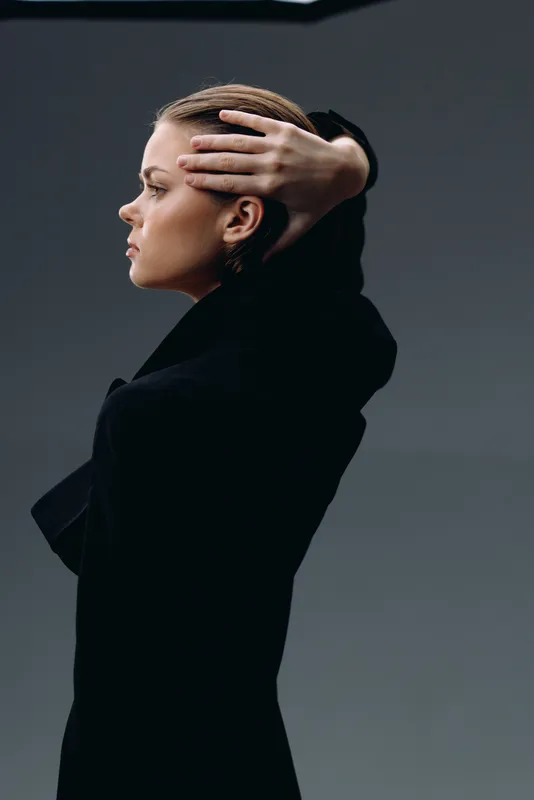 Profile of a thoughtful woman in black coat with hand on head against gray background
