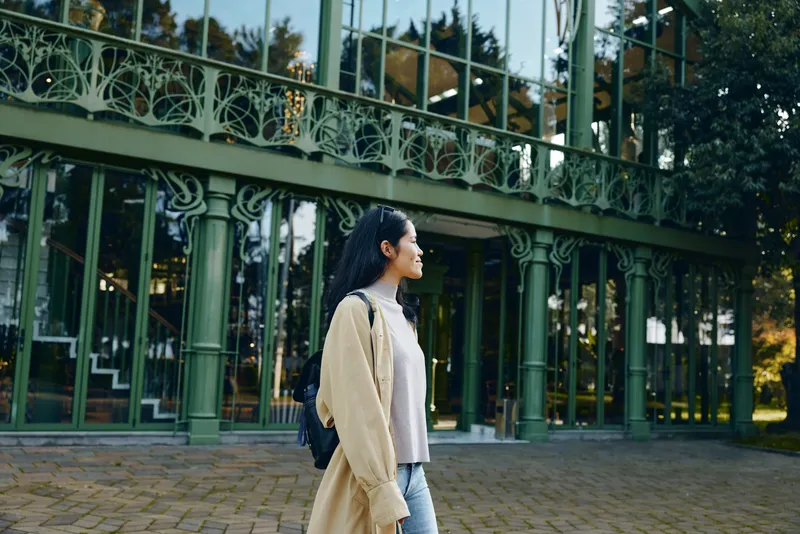 Fashion, woman with beige coat and backpack strolling beside green glass architecture in an outdoor park setting, capturing urban style and serene autumn mood