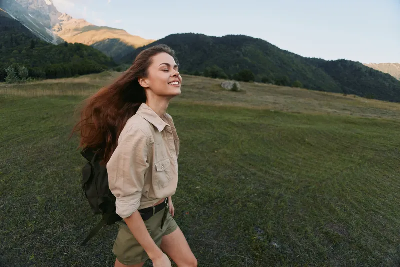 Young woman hiking outdoors with backpack, smiling with eyes closed, enjoying fresh air, casual outfit, natural landscape with mountains and grass, sunny day