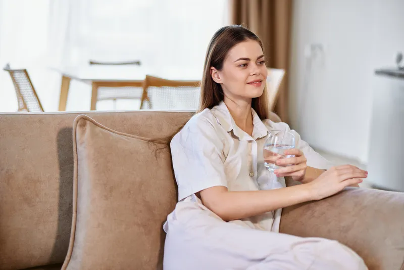 Woman on sofa sitting in a relaxed pose at home, holding a clear glass of water in the livingroom. Casual portrait of a young female enjoying calm domestic comfort and quiet hydration.