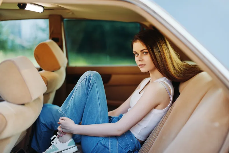 Young woman in casual outfit sitting in a car, looking thoughtful, with a serene background and soft natural lighting, conveying a sense of relaxation and freedom.