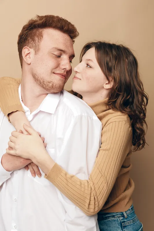 Young couple embraces tenderly, woman hugging man from behind with closed eyes. Warm beige background, casual clothing, intimate moment of affection and love between partners.