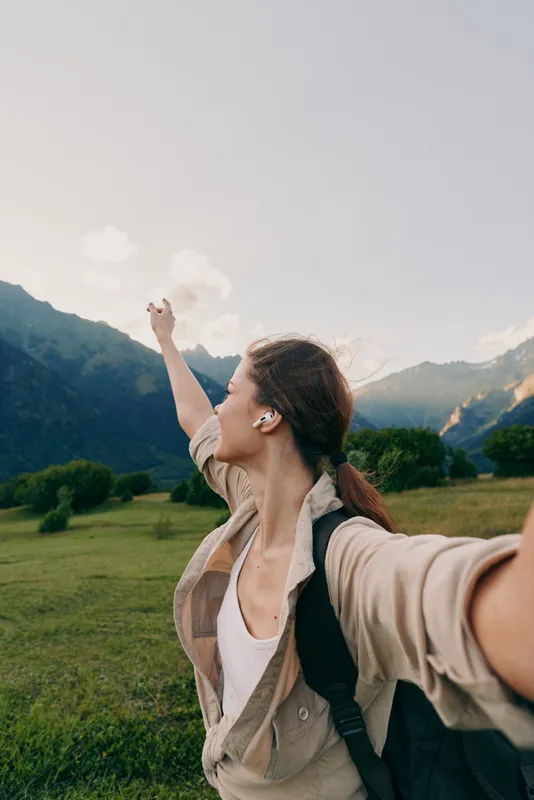 Young woman with a backpack takes a selfie in a valley as mountains rise behind, her arm extended toward the camera with a joyful breeze.