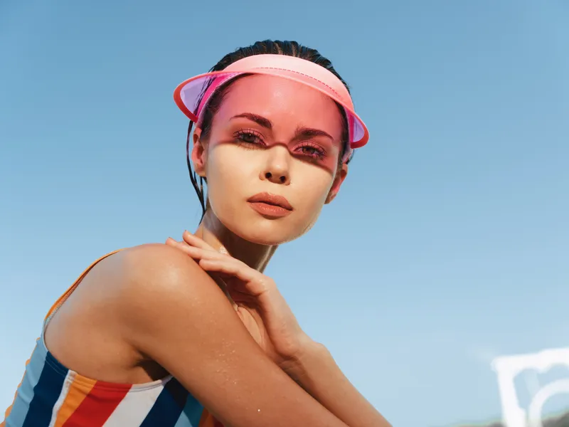 Portrait of a young woman in a colorful striped swimsuit and pink visor, sunlit skin, relaxed pose, outdoor beach scene beneath a clear blue sky.
