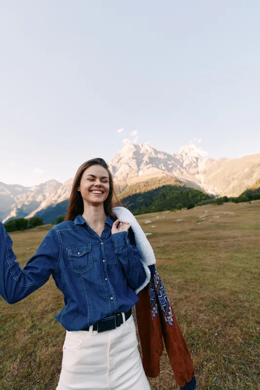 Woman smiling in denim shirt and white skirt holding a jacket on a meadow with mountains in the background. Outdoor portrait showing happiness, nature landscape, casual travel and sunshine.