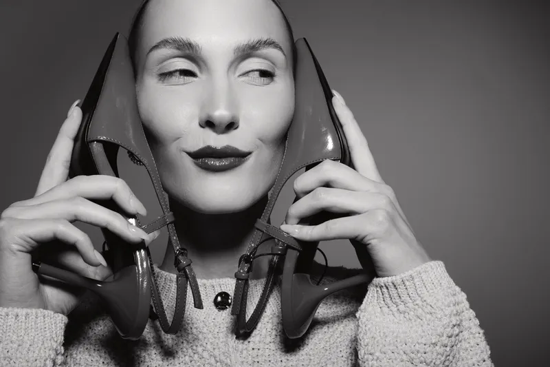 A Caucasian woman with fair skin holds red high heel shoes near her face, smiling slightly with closed eyes. Concept for fashion, style, and feminine accessories.