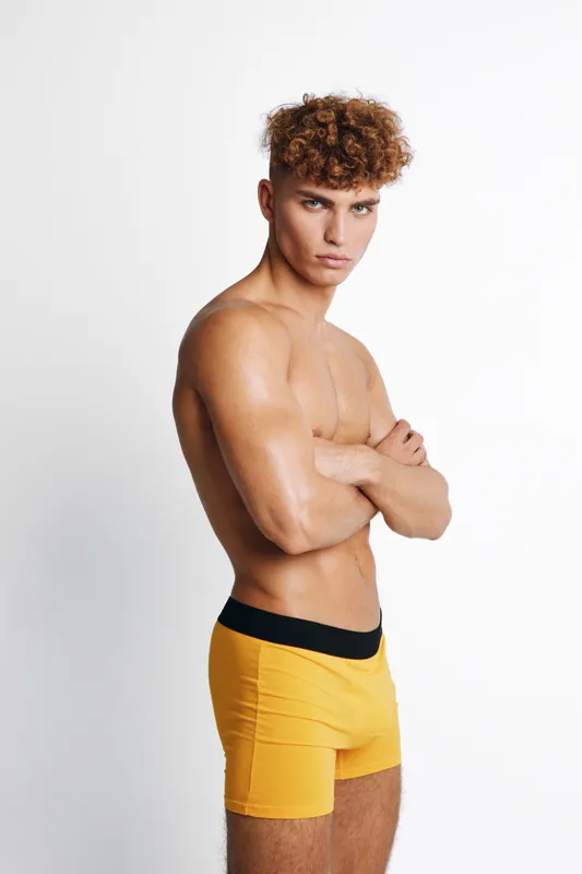 Young muscular man with curly hair posing with crossed arms wearing yellow shorts against white background. Fitness model showing strong confident posture without shirt.