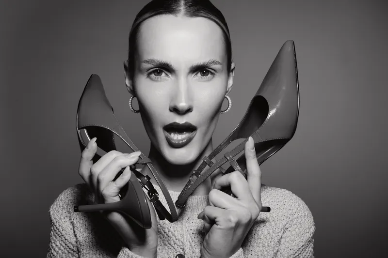 Portrait of a Caucasian woman with pale skin holding two high heel shoes near her face, expressing surprise. Studio lighting highlights facial features and shoes.
