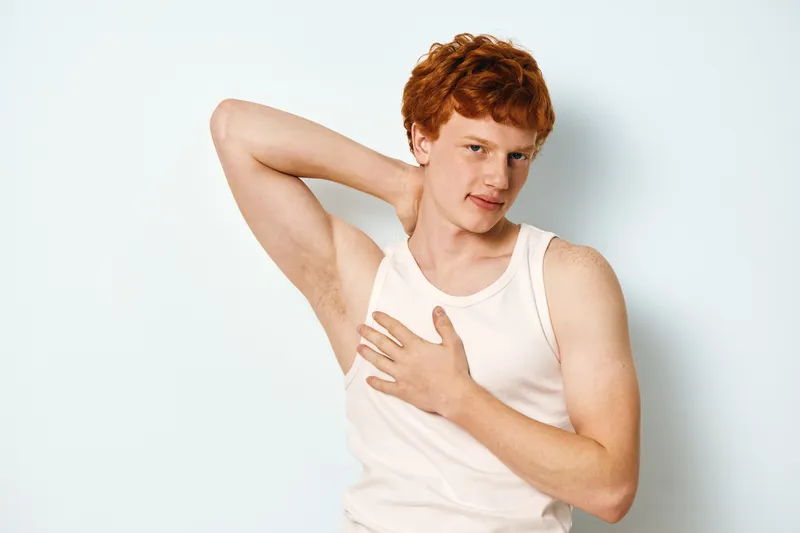 Young man with red hair wearing white tank top touching underarm with hand, posing on white background for personal hygiene or health concept portrait.