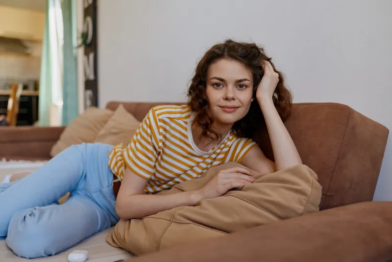 A relaxed woman lounges on a couch at home, wearing a striped shirt, light emphasizing casual comfort. A candid moment of woman lifestyle at home, self care, and repose.