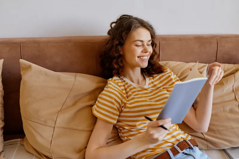 Smiling woman with curly hair wearing a striped shirt sits on a sofa and reads a notebook. Cozy home environment, casual lifestyle, and personal reflection captured in soft daylight, conveying comfort