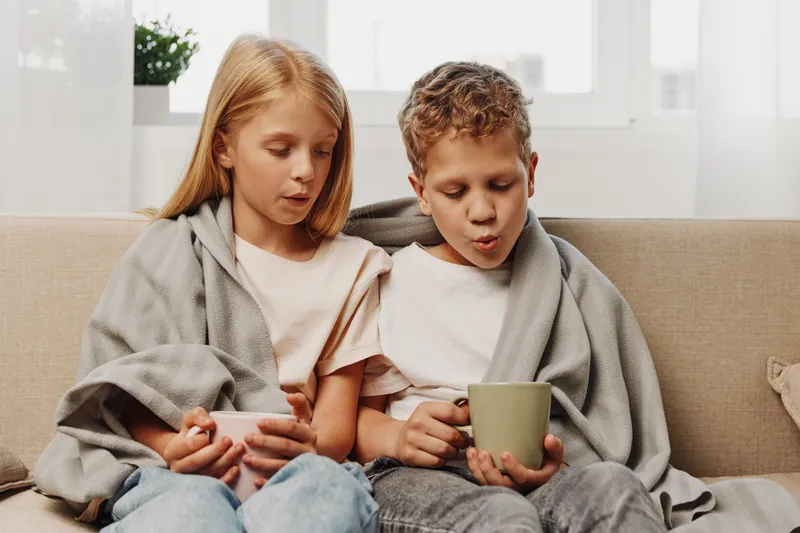 Two children wrapped in blankets sit on a couch drinking warm beverages near a bright window, showing comfort and coziness indoors.