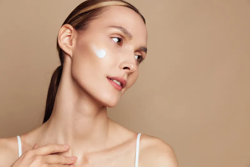 Young woman with dark hair applies white facial cream on her cheek in a soft studio, close up against a beige background, conveying skincare, self care and wellness.