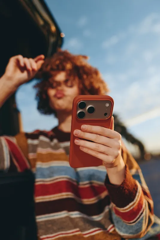 Woman selfie with smartphone in outdoors lifestyle, curly hair framing her face, striped streetwear. Another person blurred in background under bright sky, candid everyday life moment.