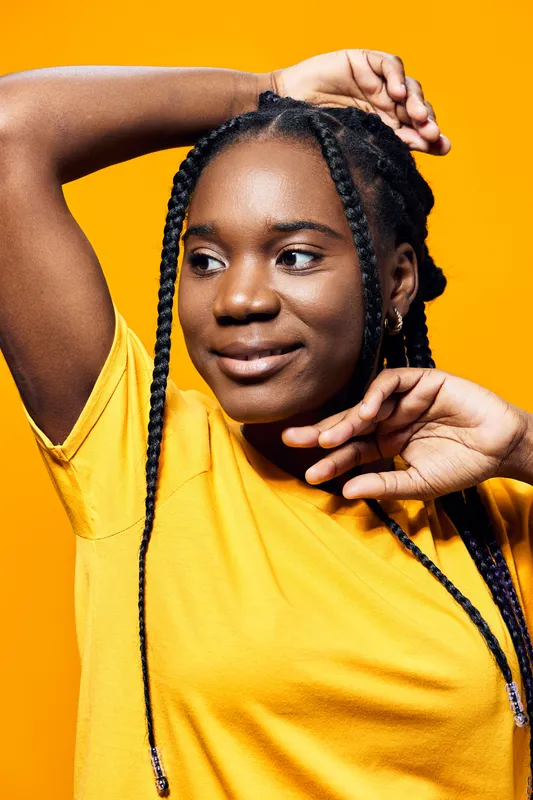 Young Black woman with braids wearing a yellow t shirt, smiling confidently against a vibrant yellow background, embodying joy and self expression