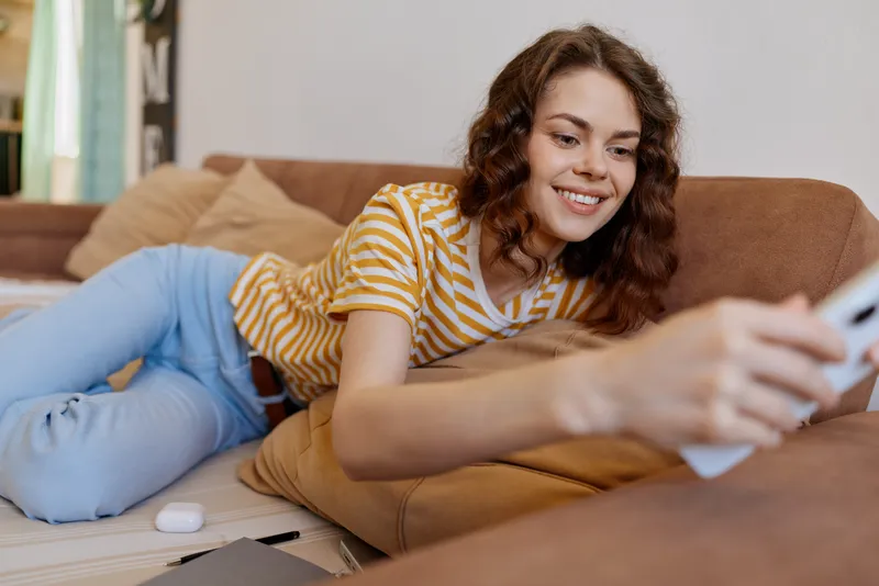 A smiling woman lounges on a sofa at home, looking at a smartphone in her hand. Bright living room, casual attire, warm light, a moment of relaxed connection.