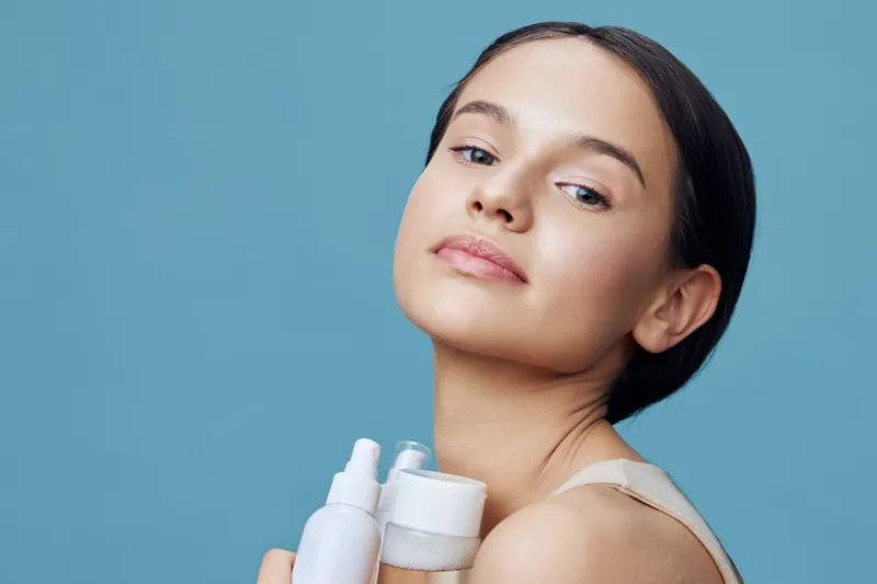Young woman with flawless skin holding skincare products against a blue background, expressing confidence and beauty Skincare, beauty, confidence, model, products, wellness