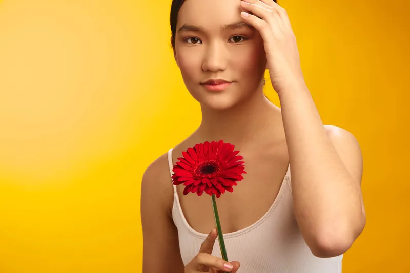 asian woman holding a red flower against a warm yellow backdrop, serene portrait with calm expression and gentle gaze, creative studio lighting, soft color palette.