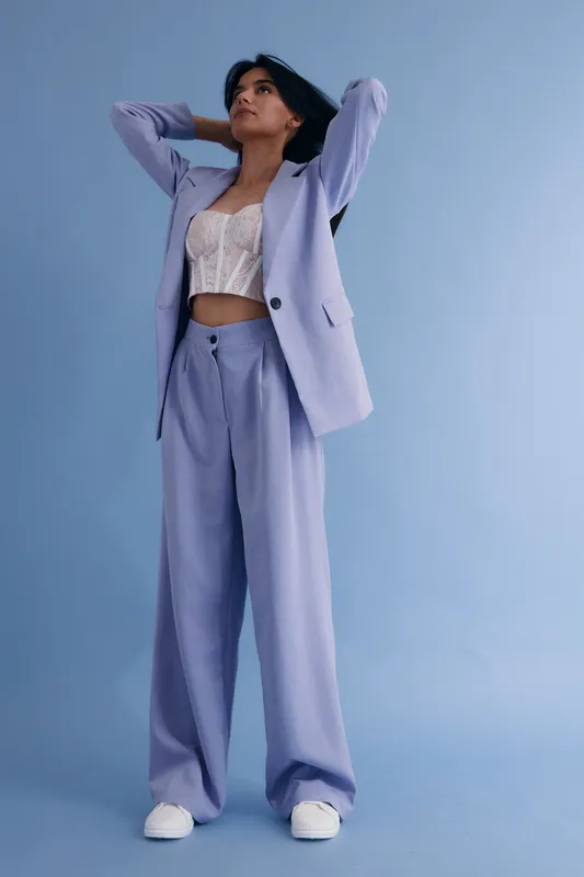 Young woman in lilac suit with lace top posing confidently against a soft blue background, showcasing modern fashion and casual elegance in studio lighting.