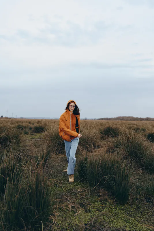 Woman lifestyle portrait, outdoors walking on a countryside path through a grassy field, nature landscape with soft light and cloudy sky, wearing brown jacket and casual jeans, calm look
