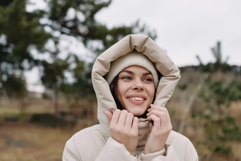 Woman outdoors in a hooded coat and parka smiles softly in a chilly landscape, a calm winter moment with cozy warmth against a windy day and natural scenery.