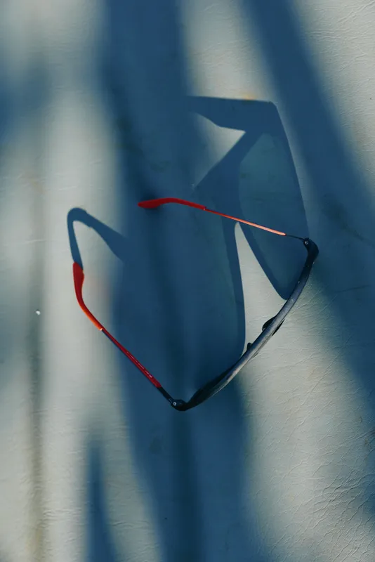 Sunglasses with red and black frames rest on a sunlit wall, casting angular shadows across the surface to evoke a casual, reflective lifestyle moment in bright light.