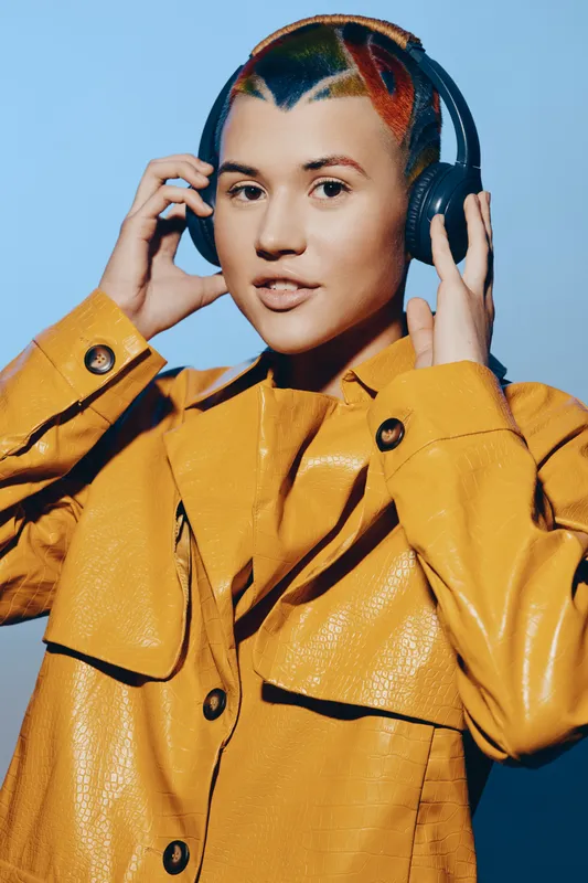 young woman with colorful hair wearing headphones and a bright yellow jacket, posing confidently against a clear blue background with modern style