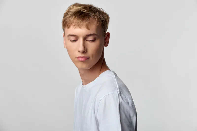 Young man in a white t shirt with a contemplative expression against a neutral backdrop, minimal studio fashion mood, clean composition, soft lighting, subtle shadows, versatile lifestyle image for