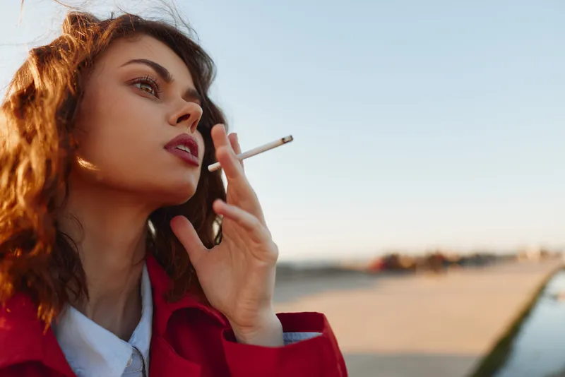 Young woman with curly brown hair smokes a cigarette by the seaside, wearing a red jacket, looking toward the horizon with a calm, rebellious mood.