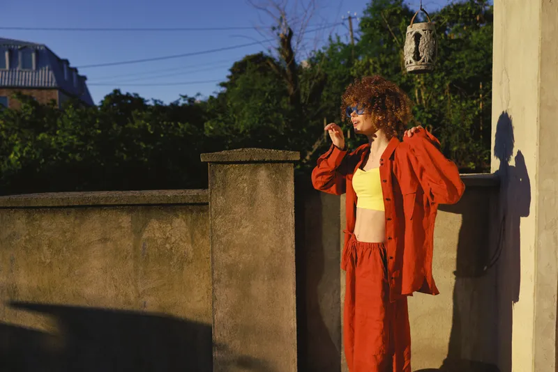 A stylish woman outdoors on a sunlit day adjusts a bold red jacket over a yellow top, embodying casual street fashion, color, confidence, and lifestyle in an urban setting