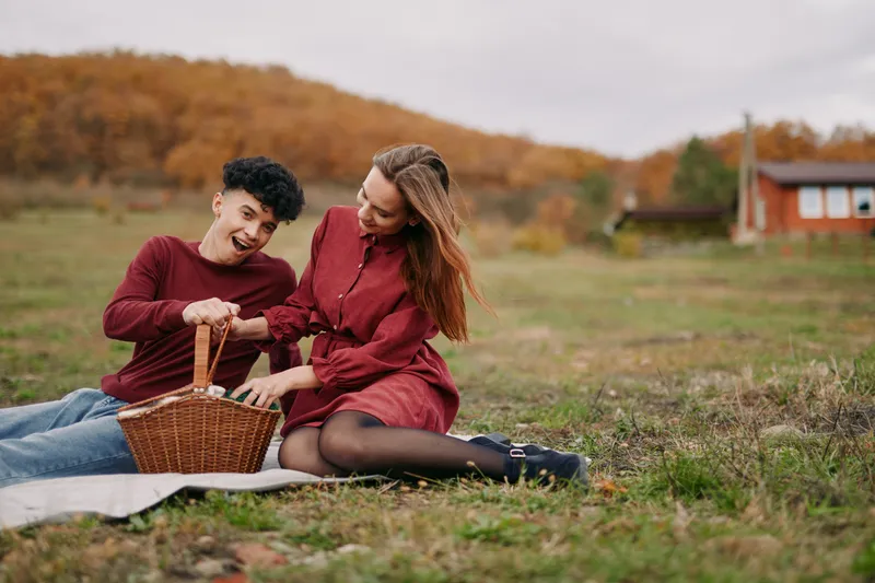 Two people share a candid autumn picnic on a sunlit field, lending authenticity to a warm, real moment as they laugh beside a wicker basket on a checkered blanket.