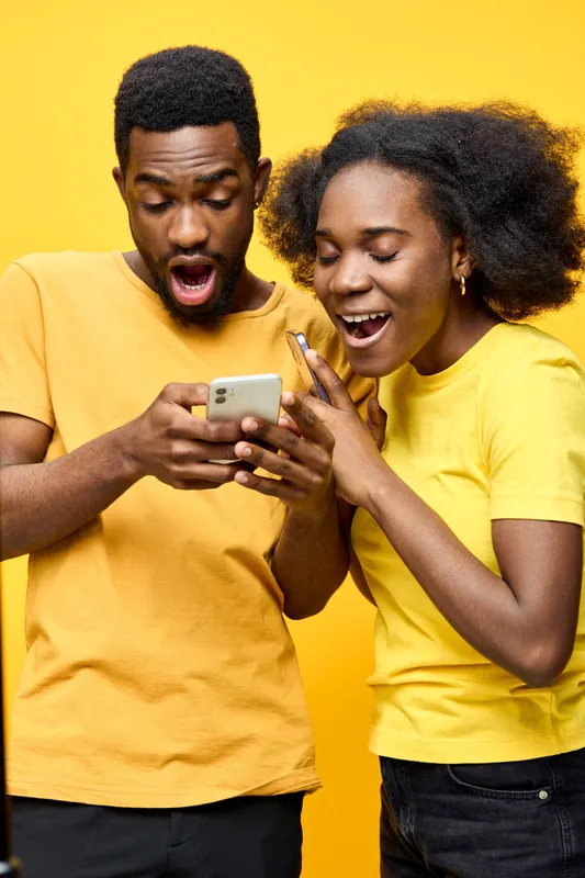 Young diverse couple expressing excitement while looking at their smartphone against a vibrant yellow background, wearing matching yellow shirts