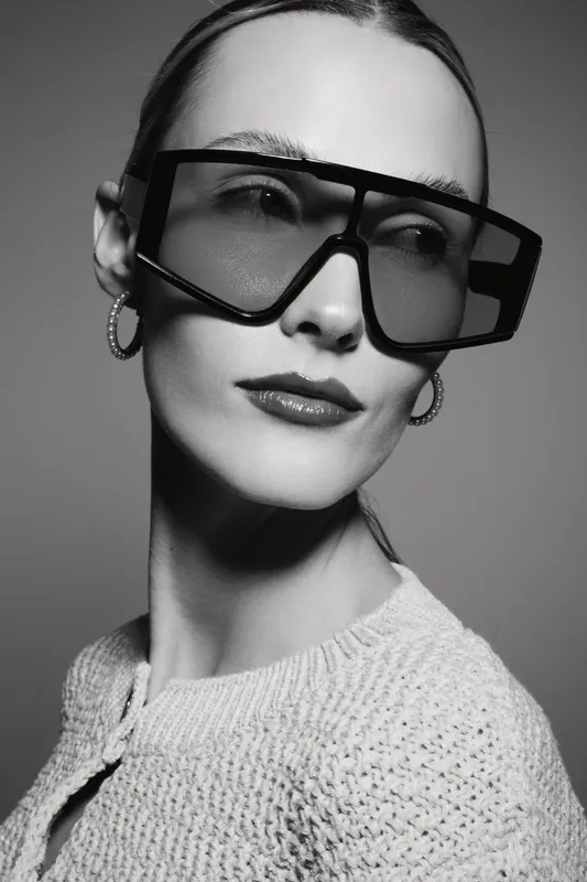 Portrait of a fair skinned Caucasian woman wearing large oversized sunglasses and hoop earrings. Studio lighting highlights her smooth skin and textured sweater.