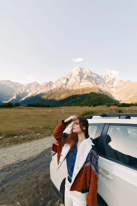 Woman with car parked by mountain landscape, enjoying travel and adventure. Casual jacket, scenic meadow and road, portrait of relaxed traveler taking in peak views.