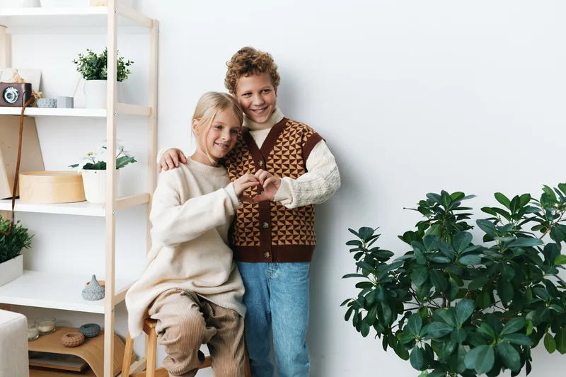Two friends embrace and smile indoors, wearing cozy sweaters, framed by a bright, tidy room with shelves, plants, soft natural light, and a calm, inviting atmosphere.