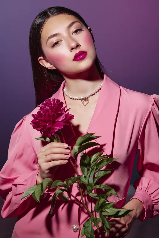 Fashion model in a blush pink blazer holds a deep pink flower with green leaves against a purple background. Her glossy hair and vibrant makeup create bold beauty creative concept.