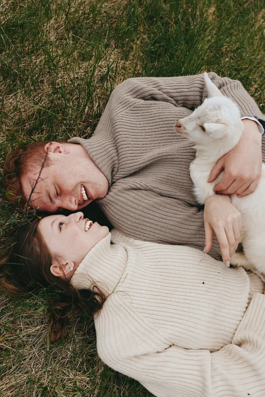 Smiling couple lying on grass wearing cozy sweaters, cuddling a goat kid in warm natural light, capturing a lifestyle moment of affection, nature, and calm outdoor comfort